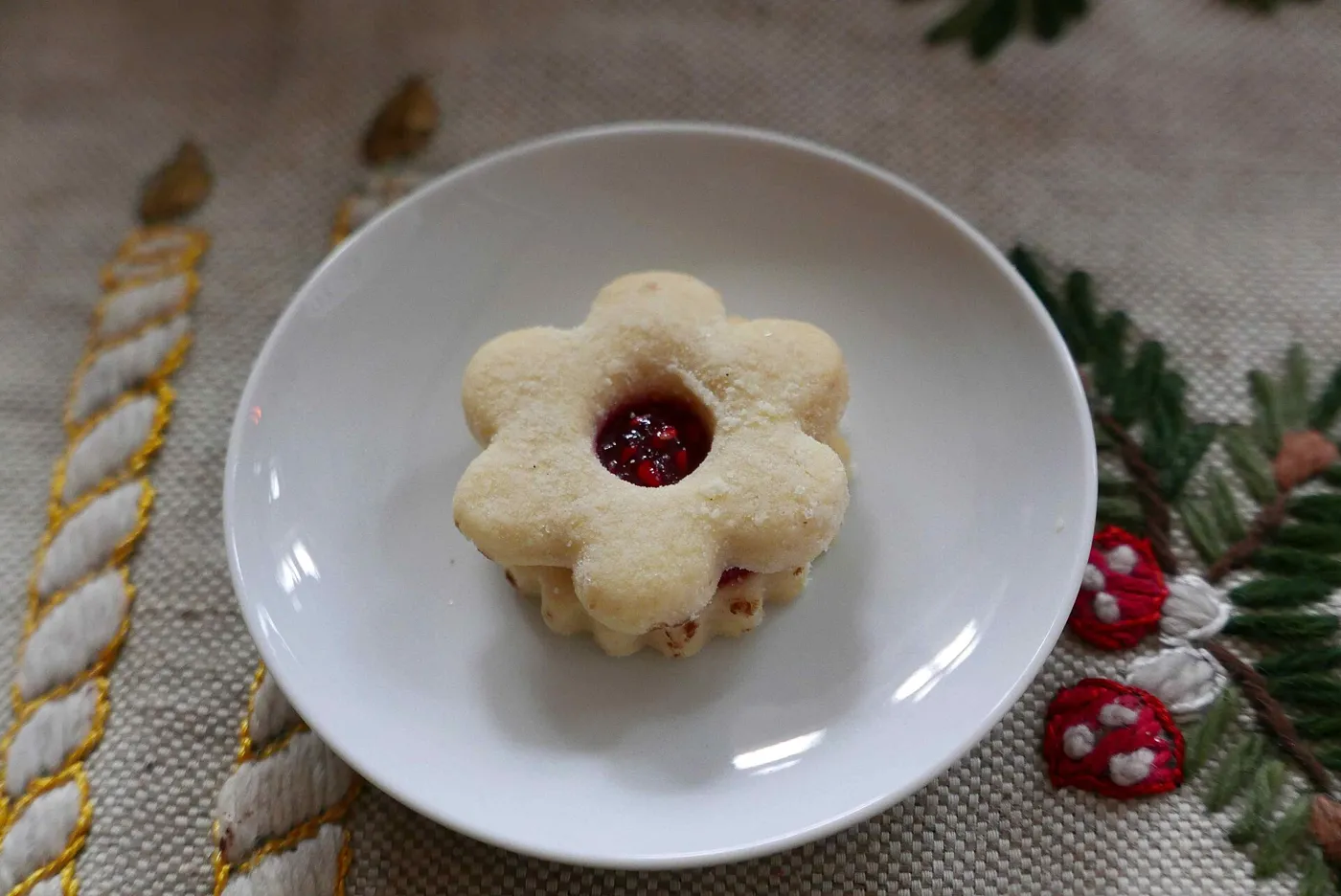 Un Spitzbub en forme de fleur, cœur de confiture de framboises, sur une nappe de Noël brodée.